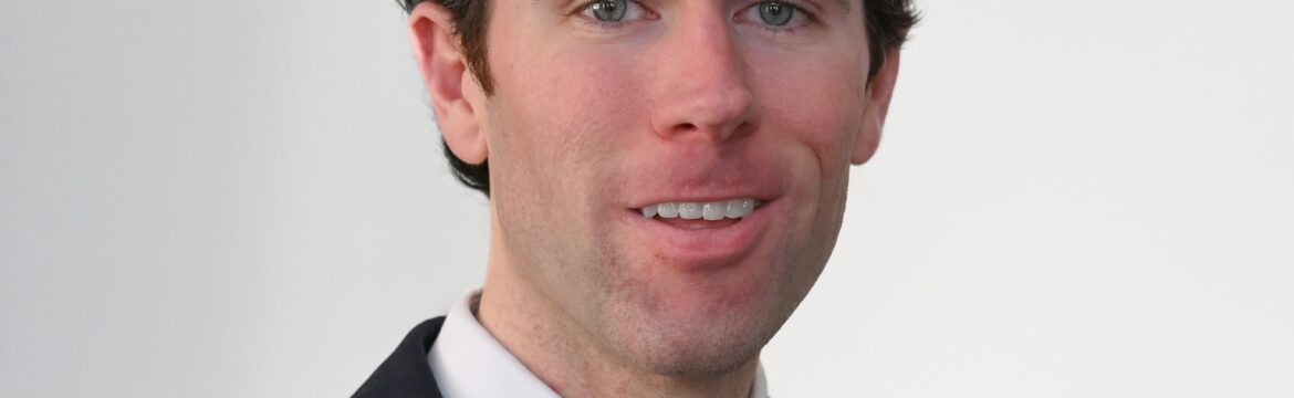 Will Simpson headshot, wearing a black jacket, white shirt, and red and navy stripe tie, white background