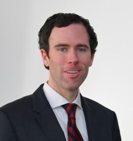 Will Simpson headshot, wearing a black jacket, white shirt, and red and navy stripe tie, white background
