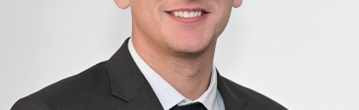 Jordan Ferguson headshot, wearing a dark grey sport coast, navy tie and white shirt, white background
