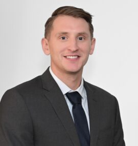 Jordan Ferguson headshot, wearing a dark grey sport coast, navy tie and white shirt, white background