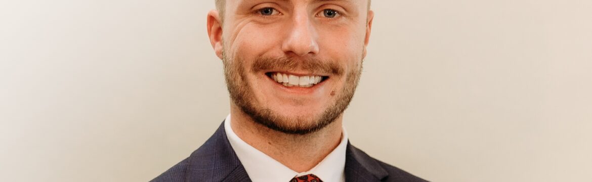 Jake Schott headshot, white shirt with red tie and black sport coat, beige background.
