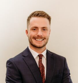 Jake Schott headshot, white shirt with red tie and black sport coat, beige background.