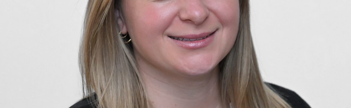 Cora Saftner headshot, wearing white shirt and black sweater, white background